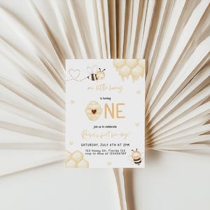 Op de afbeelding: Een witte uitnodigingskaart met een bij- en honingraatontwerp. De tekst op de kaart luidt "Mr. Little Honey wordt EEN. Vier met ons Rachels eerste bijendag! Zaterdag 4 juli om 14:00 uur. 123 Honey St, Florida 123. RSVP naar [telefoonnummer]"