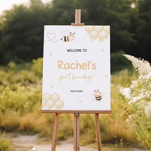 May include: Welcome to Rachel's first bee-day! A white sign with a bee and honeycomb design, the text "Welcome to Rachel's first bee-day!" and the date 04.07.2024.