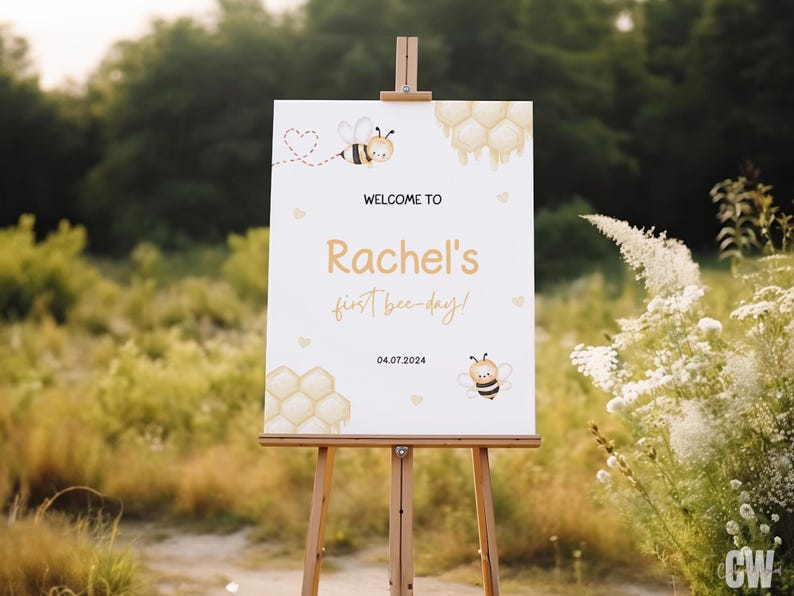 May include: Welcome to Rachel's first bee-day! A white sign with a bee and honeycomb design, the text "Welcome to Rachel's first bee-day!" and the date 04.07.2024.