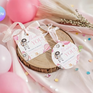 May include: Two gift tags with a space theme, featuring an astronaut, rocket, and planets in pink and white. The tags say "Thank You!" and are tied with pink ribbons. They rest on a wooden slice with pink balloons in the background.
