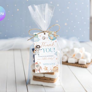 Puede incluir: Una bolsa transparente atada con hilo, que contiene malvaviscos y galletas, con una etiqueta decorativa. La etiqueta presenta un astronauta, un cohete y planetas, con el texto "Thank You!" y "So much for showering our little baby!"