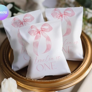 May include: Three white party favor bags with pink watercolor bows and ribbons. The bags have the text "Amelia is ONE" and "Lia is ONE" in pink script. The bags are on a gold-colored tray.