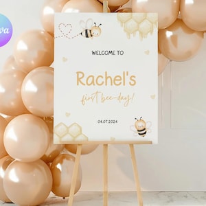 May include: Welcome to Rachel's first bee-day! A white sign with a bee and honeycomb design, the text "Welcome to Rachel's first bee-day!" and the date 04.07.2024. The sign is on a wooden easel and surrounded by beige balloons.