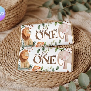 May include: Two chocolate bar wrappers with a jungle theme. Each wrapper has a lion, monkey, and elephant illustration with the words "wild ONE" in brown. The wrappers are on a woven placemat.