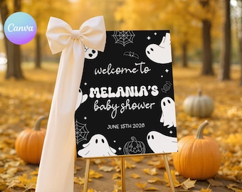 May include: A black sign with white ghost and spiderweb illustrations, welcoming guests to Melania's baby shower on June 15th, 2026. The sign is propped on a gold easel and adorned with a cream-colored bow. Pumpkins and fall foliage surround the sign.