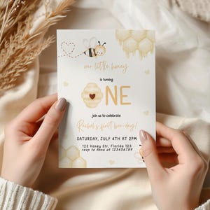 Op de afbeelding: Een witte uitnodigingskaart met een bij- en honingraatontwerp. De tekst luidt "Our little honey is turning ONE. Join us to celebrate Rachel's first bee-day! Saturday, July 4th at 2PM. 123 Honey Str, Florida 123. rsvp to Anna at 123456789"