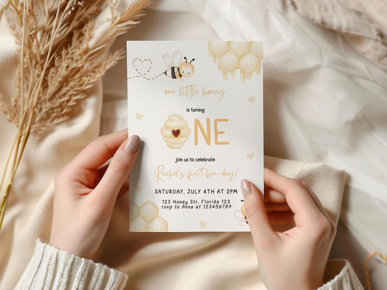 Op de afbeelding: Een witte uitnodigingskaart met een bij- en honingraatontwerp. De tekst luidt "Our little honey is turning ONE. Join us to celebrate Rachel's first bee-day! Saturday, July 4th at 2PM. 123 Honey Str, Florida 123. rsvp to Anna at 123456789"