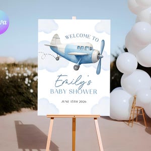 May include: A baby shower sign on an easel features a watercolour illustration of a blue and white aeroplane with the text "Welcome to Emily's Baby Shower" and the date June 15th, 2026. White balloons are in the background.