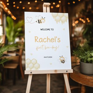 May include: White welcome sign with a bee and honeycomb design. The sign says "Welcome to Rachel's first bee-day! 04.07.2024".