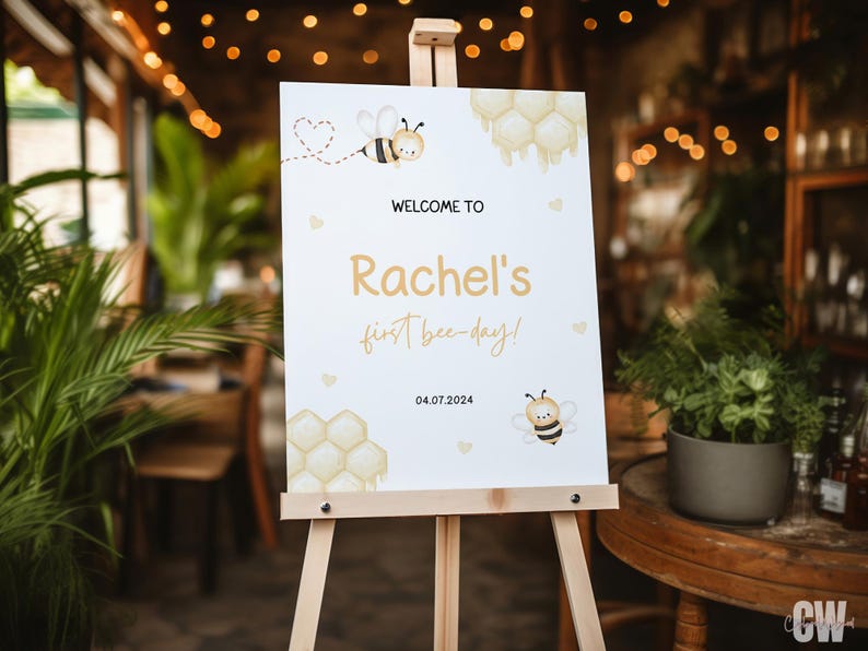May include: White welcome sign with a bee and honeycomb design. The sign says "Welcome to Rachel's first bee-day! 04.07.2024".