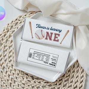 May include: Two chocolate bars with baseball designs. The top bar says "Kevin is turning ONE" with a baseball and bats. The bottom bar has a nutrition facts label. The bars are on a white plate.