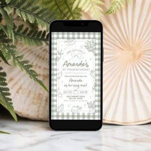 May include: A smartphone displays a digital invitation for Amanda's 1st Farmers Market, with a green gingham background. The invitation features hand-drawn illustrations of produce and the text "Amanda is turning one!" The date is July 5th.