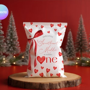 Może przedstawiać: Biała torba na upominki z czerwonymi sercami i czerwoną wstążką. Na torbie widnieje napis "Our Little Sweetheart Maddie is turning one". Torba leży na drewnianej powierzchni z choinkami w tle.