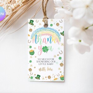 Puede incluir: Una etiqueta de regalo rectangular con un arcoíris, tréboles y las palabras "gracias". La etiqueta también incluye el texto "¡Tanto por bañar a nuestro pequeño bebé! con amor". Se adjunta una cuerda marrón.
