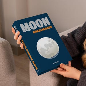 May include: A book titled "MOON DREAMSCAPE" with a full moon graphic on the cover. The book has a dark blue cover with white and orange text. The book is held by a person.