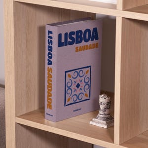 May include: A book titled "LISBOA SAUDADE" with a decorative tile design on the cover, displayed on a wooden shelf. The book's spine reads "LISBOA SAUDADE". A small, white decorative statue sits next to the book. The shelf is part of a wooden bookcase.