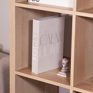 May include: A light-colored wooden bookshelf with a book titled "CALM HOME" and a small decorative statue. The book is light gray with the title in large, light gray letters. The bookshelf has multiple shelves.