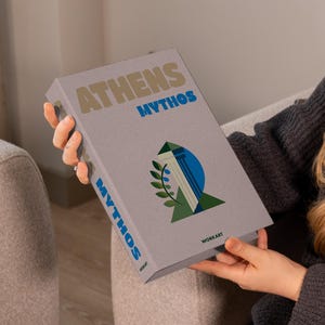May include: A person holds a book titled "Athens Mythos". The book has a grey cover with the title in beige and blue, and a graphic of a column, a blue circle, and a green leaf. The book's spine also reads "Mythos".