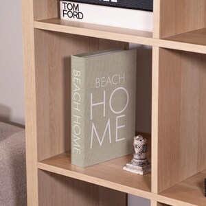 K&ouml;nnte beinhalten: Ein helles B&uuml;cherregal mit mehreren F&auml;chern. Ein Buch mit dem Titel "BEACH HOME" steht auf einem Regal. Eine kleine dekorative Statue und ein Buch mit dem Titel "TOM FORD" sind ebenfalls zu sehen.