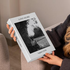 May include: A rectangular white photo album with a black and white photograph of a couple sitting under a tree on the cover. The text LOVE STORY is printed at the top and on the spine, with the date August 1, 2026, printed at the bottom.