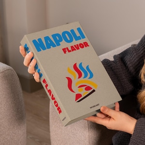 May include: A rectangular book with the words "NAPOLI FLAVOR" in blue and red. The cover features a colorful graphic of a pizza and flames. The book is held by a person wearing a gray sweater.