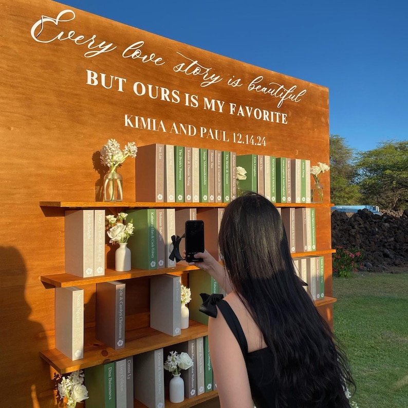 May include: A wooden wedding display with the text "Every love story is beautiful BUT OURS IS MY FAVORITE KIMIA AND PAUL 12.14.24". The display features shelves with books and small vases of white flowers. A person is taking a photo of the display.