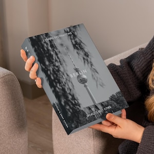 Op de afbeelding: Een boek getiteld "Berlin" uit de serie "World's Best Cities". De omslag toont een grijswaardenafbeelding van de Berlijnse televisietoren. Het boek wordt vastgehouden door een persoon die een donkergrijze trui draagt.