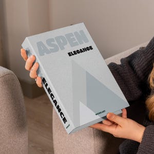May include: A book titled "ASPEN ELEGANCE" is held by a person. The book has a light gray cover with the title in gray and black text. A geometric design in shades of gray is on the cover. The word "ELEGANCE" is also printed vertically on the spine.