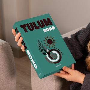 May include: A teal book titled "Tulum Boho" with a sun, leaf, and spiral design. The book is held by a person wearing a gray sweater. The book's cover has the word "WORKART" at the bottom.