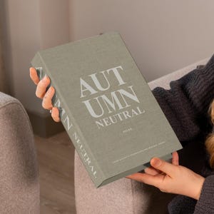 May include: A light grey decorative book with the title "AUTUMN NEUTRAL" in white lettering. The book is held by a person wearing a dark grey sweater. The book's spine also displays the words "AUTUMN NEUTRAL". The book is a decor item.