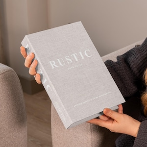 Könnte beinhalten: Ein hellgraues Buch mit dem Titel "RUSTIC" in weißen Großbuchstaben. Das Buch hat einen strukturierten Einband und wird von einer Person gehalten, die einen grauen Pullover trägt. Das Thema des Buches ist Inneneinrichtung.