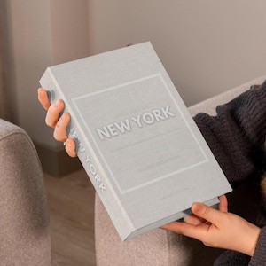以下が含まれることがあります： 人が持っている「Little Book of New York」というタイトルの薄い灰色の本。本は長方形で、タイトルは白い浮き出し文字です。本の表紙は質感があります。
