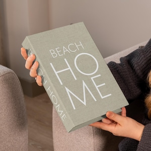K&ouml;nnte beinhalten: Ein salbeigr&uuml;nes Buch mit dem Titel "BEACH HOME" in wei&szlig;en Gro&szlig;buchstaben. Das Buch wird von einer Person gehalten, die einen dunkelgrauen Pullover tr&auml;gt. Der Einband des Buches hat eine strukturierte Oberfl&auml;che, und die Person sitzt auf einem hellen Sofa.