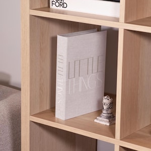 Puede incluir: Una estantería de madera clara con múltiples estantes. Un libro titulado "LITTLE THINGS" se exhibe en un estante, junto con una pequeña estatua decorativa. Otro libro es visible en el estante superior.