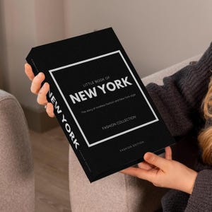 May include: A black book titled "Little Book of New York" with white text and a white border. The book's cover includes the text "Fashion Collection" and "Fashion Edition". The book is held in someone's hands.