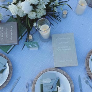 May include: A wedding table setting with white roses, candles, and place settings. The table is set with woven placemats, white plates, and silverware. Books with the text "Every love story is beautiful, but ours is my favorite" are placed on the table.