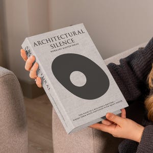 Op de afbeelding: Een boek getiteld "Architectural Silence" uit de Workart Master Series. Het boek heeft een lichtgrijze omslag met een groot zwart ovaal ontwerp en witte tekst. De tekst op de omslag bevat de titel en de serienaam. Het boek wordt vastgehouden.