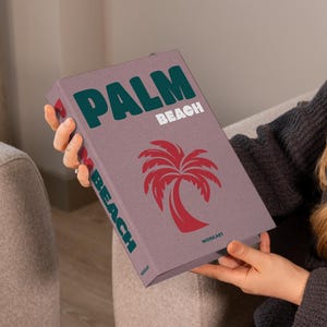 May include: A book with the words "PALM BEACH" in teal and white, and a red palm tree graphic. The book is held by a person wearing a gray sweater. The book's cover is a muted purple color.