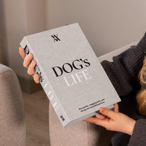 May include: A light grey book titled "DOG'S LIFE" is held. The book has a textured cover and features the text "Paw prints, wagging tails, and the joy of unconditional love." The book's cover also has a small logo.