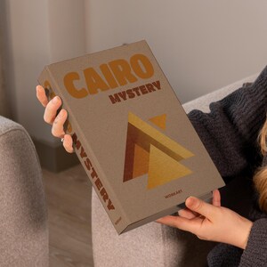 May include: A hardcover book titled "CAIRO MYSTERY" with a geometric design in shades of brown and gold. The book is held by a person wearing a gray sweater. The book's cover has the word "MYSTERY" printed vertically.