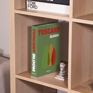 May include: A light wood bookcase with multiple shelves. A green book titled "TUSCANY BLOOM" with colorful abstract shapes is on one shelf. A small decorative statue and a book titled "TOM FORD" are also visible.