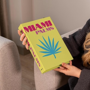 May include: A book titled "MIAMI PALMS" with a light green cover. The cover features the title in pink, a blue palm leaf illustration, and the title repeated on the spine. The book is held by a person.