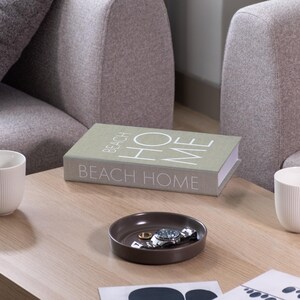 K&ouml;nnte beinhalten: Ein Couchtisch mit einem Buch mit der Aufschrift "BEACH HOME" und zwei wei&szlig;en Tassen. Eine kleine braune Schale enth&auml;lt eine Uhr und Ringe. Der Tisch steht vor zwei grauen Polstersesseln.