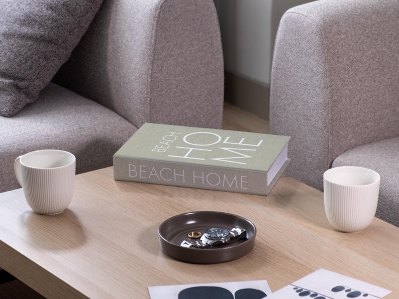 K&ouml;nnte beinhalten: Ein Couchtisch mit einem Buch mit der Aufschrift "BEACH HOME" und zwei wei&szlig;en Tassen. Eine kleine braune Schale enth&auml;lt eine Uhr und Ringe. Der Tisch steht vor zwei grauen Polstersesseln.