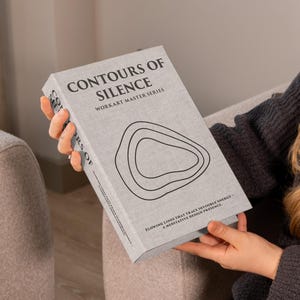 Könnte beinhalten: Ein Buch mit dem Titel "CONTOURS OF SILENCE" aus der WORKART MASTER SERIES wird gehalten. Der Einband ist hellgrau mit schwarzem Text und einer Grafik konzentrischer Formen. Der Buchrücken ist sichtbar, und der Text "ARS OF" ist teilweise sichtbar.