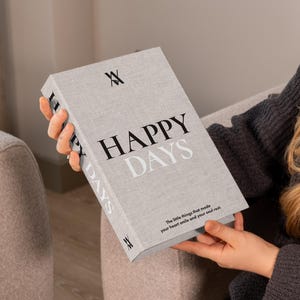 May include: A light gray book titled "HAPPY DAYS" with black and light gray text. The book has the text "The little things that made your heart smile and your soul rest." The book is held by a person.