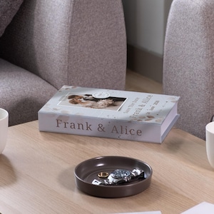 May include: A coffee table setting featuring a decorative book with the names "Frank & Alice" and a photo, a small brown tray with a watch and ring, and two white ribbed mugs. The scene is set in a living room.