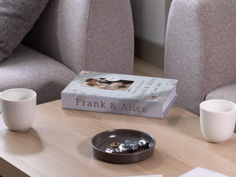May include: A coffee table setting featuring a decorative book with the names "Frank & Alice" and a photo, a small brown tray with a watch and ring, and two white ribbed mugs. The scene is set in a living room.