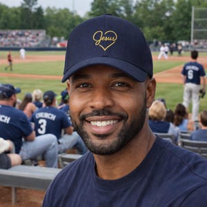 Könnte beinhalten: Marineblaue Baseballkappe mit goldenem Herz und dem Wort "Jesus". Die Kappe wird von einer lächelnden Person getragen. Die Person trägt ein marineblaues T-Shirt. Im Hintergrund ist ein Baseballspiel zu sehen.