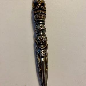 May include: A silver dagger with detailed carvings. The handle features a skull design, ornate patterns, and a dragon's head. The blade is pointed and metallic. The dagger's design suggests a ceremonial or decorative purpose.